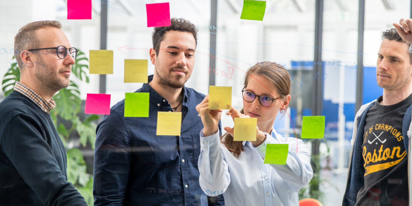 Vier Personen stehen vor einem transparenten Whiteboard. Auf dem Board befinden sich mehrere farbige Post-Its, welche an farbigen Linien angebracht sind. Die weibliche Person beschriftet eines der Post-Its. Neben ihr stehen drei männliche Peronen, welche auf das Post-It schauen.