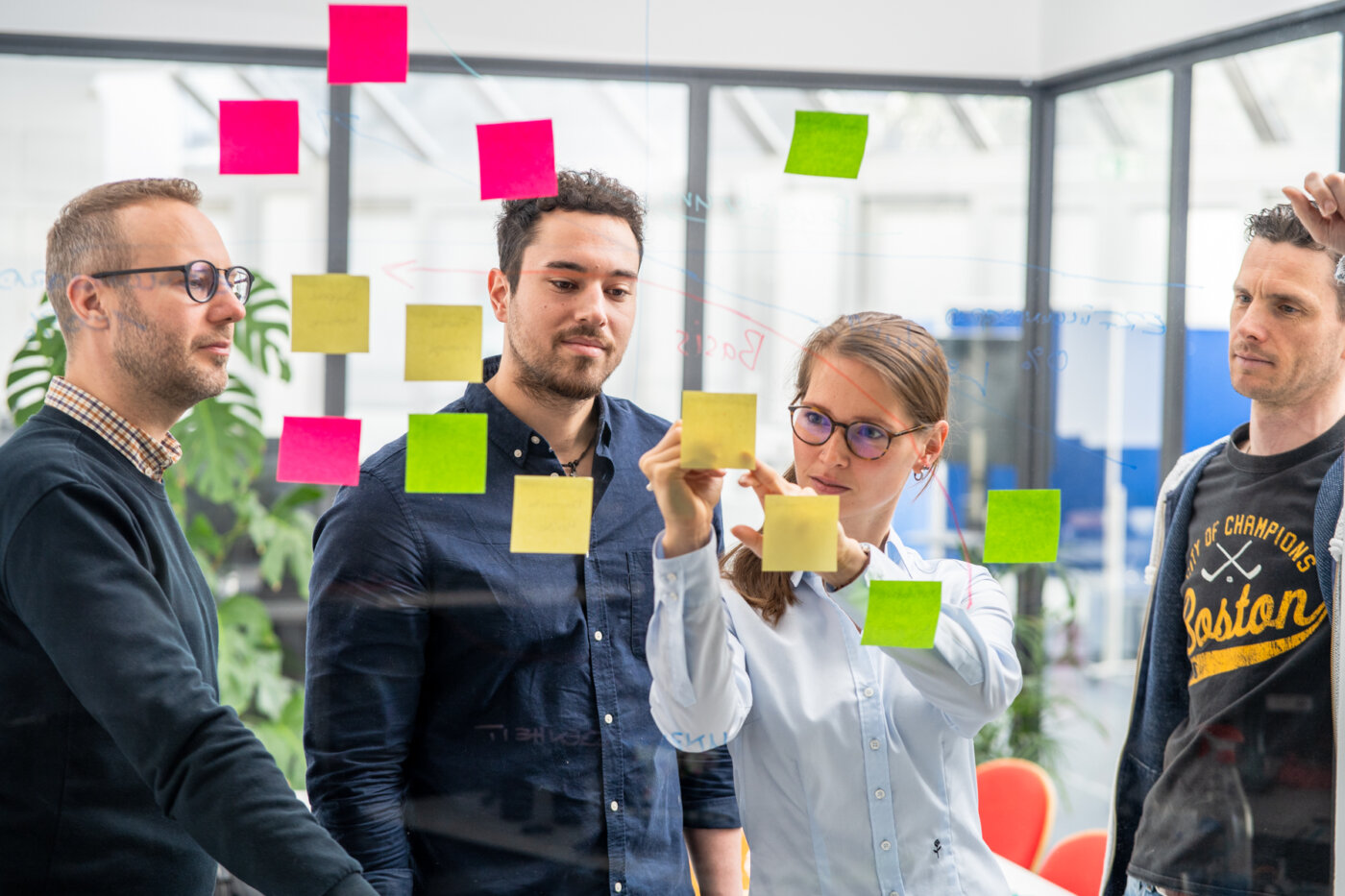 Vier Personen stehen vor einem transparenten Whiteboard. Auf dem Board befinden sich mehrere farbige Post-Its, welche an farbigen Linien angebracht sind. Die weibliche Person beschriftet eines der Post-Its. Neben ihr stehen drei männliche Peronen, welche auf das Post-It schauen.