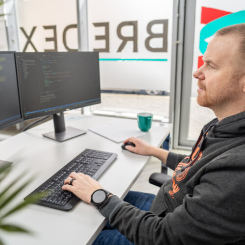 Ein weißer Mann mit kurzem braunen Haaren und einem Bart sitzt an einem Schreibtisch vor zwei Monitoren. Auf dem Bildschirm sind Codezeilen zu sehen.